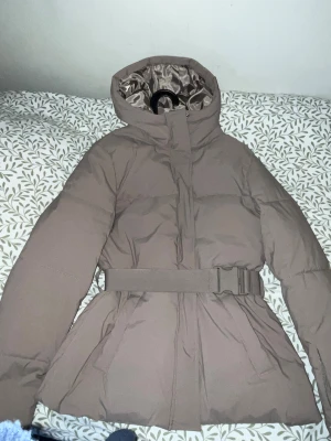 Beige pufferjacka med bälte och huva - Snygg beige pufferjacka med huva och markerad midja tack vare ett brett bälte med spänne. Jackan har långa ärmar, dragkedja framtill och två fickor. Perfekt för kalla dagar när du vill hålla stilen och värmen.