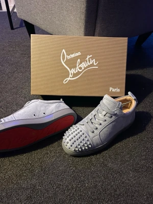 Grå Christian Louboutin sneakers med nitar - Säljer ett par turkos typ slash grå  sneakers från Christian Louboutin med ikoniska röda sulor och coola nitar på tån. Skorna har snörning och är tillverkade i mocka med rund tå och platt sula. Perfekt för dig som vill sticka ut med lyxig streetstyle.