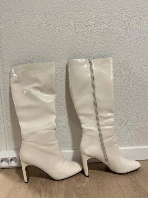 Vita höga stövlar med klack - Säljer ett par vita, glansiga stövlar med spetsig tå och hög blockklack. Stövlarna har dragkedja på insidan och är tillverkade i syntetmaterial med en slät finish. Perfekt för dig som vill sticka ut med ett statement-plagg.