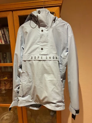 Ljusblå skidjacka från Dope Snow - Säljer en ljusblå skidjacka från Dope Snow med stor huva, dragkedja i sidan och tryckknappar framtill. Jackan har en stor ficka på magen och justerbara muddar med kardborre. Perfekt för dig som gillar streetwear och snygg design. Mycket fint skick 