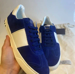 Royal blue Valentino Flycrew - Säljer ett par Valentino Flycrew sneakers i Royal blue mocka med vita skinnpaneler och coola silvriga nitar på hälen. Skorna har snörning, rund tå och en vit platt sula med mönstrad undersida. Perfekt för dig som gillar exklusiv streetstyle och snygga detaljer. En nit på hälen är tyvärr av men det är inget som syns.