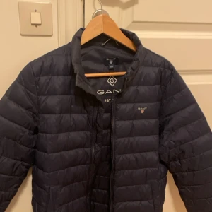 Mörkblå pufferjacka från GANT - Snygg mörkblå pufferjacka från GANT med diskret logga på bröstet och klassisk ståkrage. Jackan har horisontella quiltade sömmar och dragkedja framtill. Perfekt för kyliga dagar och har en stilren look som funkar till det mesta.