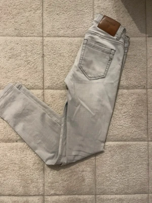 Ljusgrå jeans från Dondup - Säljer ett par ljusgrå jeans från Dondup med klassisk femficksdesign och snygga kontrastsömmar. Jeansen har en stilren look med läderpatch bak i midjan och är tillverkade i mjukt bomullsmaterial. Perfekta för dig som gillar en clean och modern stil.