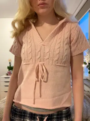Supersöt ljusrosa stickad tröja med korta ärmar och v-ringad krage. Tröjan har kabelstickat mönster upptill och en snygg snörning framtill som ger en chill vibe. Perfekt för dig som gillar mjuka pasteller och vill ha något som sticker ut lite extra.