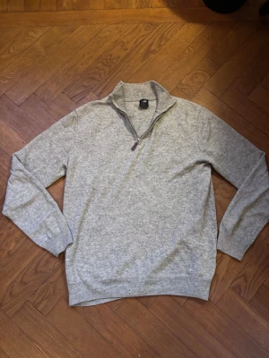 Grå halvziptröja i ull från Bvaldi - Gråmelerad stickad tröja från Bvaldi i ullblandning. Halvzip med dragkedja och läderdetalj, broderad logga på bröstet och ribbade muddar. Perfekt för lager-på-lager och chill höst- och vinterstil.