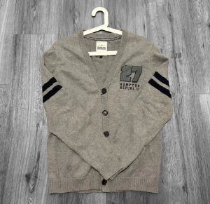 Grå kofta från Hampton Republic - Grå kofta från Hampton Republic med svarta ränder på ärmarna och broderad logga med siffran 27 på bröstet. V-ringad modell med knappar framtill och ribbade muddar. Perfekt för dig som gillar klassisk och sportig stil.