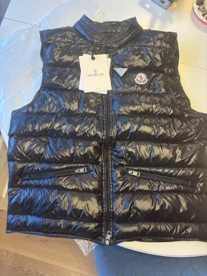 Svart dunväst från Moncler - Snygg svart dunväst från Moncler med glansig finish och quiltad design. Västen har två praktiska fickor med dragkedja, hög krage och klassisk Moncler-logga på bröstet. Kommer med originalpåse och taggar. Storlek 3 (m-l) 