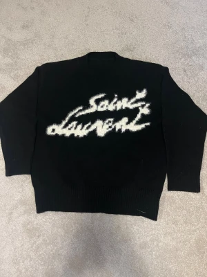 Svart Saint Laurent stickad tröja - Svart stickad tröja från Saint Laurent med vit, fluffig logotyp framtill. Tar också emot byten mot en saint Laurent hoodie! Hör av er vid minsta lilla fundering!