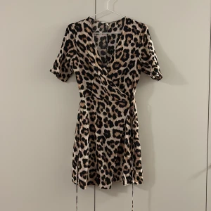 Leopardmönstrad omlottklänning H&M - Kort omlottklänning från H&M med leopardmönster i beige, svart och bruna toner. Klänningen har korta ärmar, knytband i midjan och v-ringad hals. Perfekt för dig som vill sticka ut med ett djurmönster och en feminin siluett.