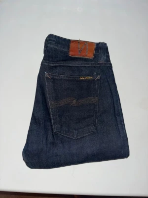 Nudie Jeans - Mörkblå Skinny Lin jeans från Nudie Jeans med smal passform och klassisk femficksdesign. Snygga kontrastsömmar och läderpatch baktill. Midja 35cm, längd 107cm. JAG SKICKAR EJ FLER BILDER NÄR JAG BÄR JEANSEN! för storleksguide hänvisar jag till måtten och för bättre inblick i passformen rekommenderar jag att googla på modellnamnet. kolla måtten noggrant :) 75
