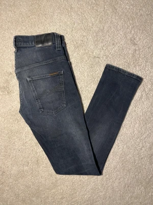 Nudie Jeans Tape Ted blåsvarta jeans - Snygga Tape Ted jeans från Nudie Jeans i blåsvart tvätt med klassisk femficksdesign. Modellen har smal passform med tapered ben och är tillverkad av 99% ekologisk bomull och 1% elastan för extra stretch. Perfekt för dig som gillar stilrena och moderna jeans.