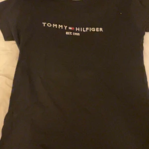 Svart Tommy Hilfiger t-shirt L - Svart t-shirt från Tommy Hilfiger i storlek L med klassisk logotyp och text 'EST. 1985' på bröstet. Tillverkad av 52% återvunnen bomull, vilket gör den både mjuk och hållbar. Enkel och stilren design med korta ärmar och rund hals.