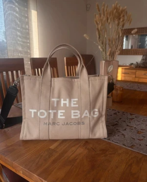 Beige tote bag från Marc Jacobs - Säljer en beige tote bag från Marc Jacobs med tydlig logga framtill. Väskan är i kraftig bomullscanvas och har både handtag och avtagbar axelrem. Perfekt storlek för vardagens alla prylar och stilren design som passar till det mesta.