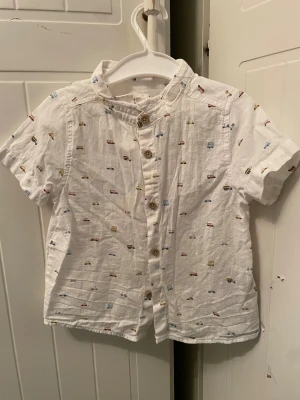 Vit kortärmad skjorta med biltryck - Söt vit skjorta med korta ärmar och små färgglada bilar tryckta över hela tyget. Skjortan har knappar framtill och en klassisk krage. Perfekt för dig som gillar lekfulla detaljer och en avslappnad stil.