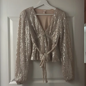 Glittrig blus med knyt från Nelly🩷 - Superfin glittrig blus i champagnefärg med v-ringning och knyt i midjan. Perfekt för nyår! Blusen har paljetter över hela tyget och ger en lyxig känsla.