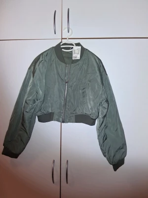 Grön croppad bomberjacka H&M - Snygg croppad bomberjacka från H&M i mörkgrön färg. Jackan har långärmade ärmar, ribbade muddar och dragkedja framtill. Tillverkad i polyester med klassisk bomberkrage och stilren look. Perfekt för dig som gillar streetstyle. Aldrig använd! 