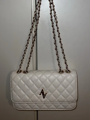 Vit quiltad axelväska från NA-KD - Snygg vit axelväska från NA-KD med quiltat mönster och guldfärgade kedjor. Väskan har ett stilrent AV-emblem framtill och flera fack inuti, inklusive ett med dragkedja. Perfekt storlek för det viktigaste och ger en lyxig vibe till din outfit. Tips: Tvätta innan användning eftersom den är lite gråaktig!