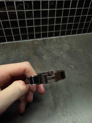 Svart Hermès armband  - Säljer ett snyggt Hermès H-armband i silverfärgad metall med svarta detaljer. Armbandet har den klassiska H-loggan framtill och en stilren, rundad form. Perfekt accessoar för dig som gillar lyxiga och tidlösa smycken.