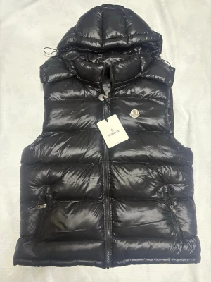 Moncler Väst - Säljer en svart, glansig dunväst från Moncler. Västen har huva (avtagbar), två fickor med dragkedja och klassisk Moncler-logga på bröstet. Insidan har ett coolt serietryck och etikett. Perfekt för lager-på-lager och streetstyle. Det finns en liten defekt inuti vänstra ficka, inget som stör funktionen dock 😊