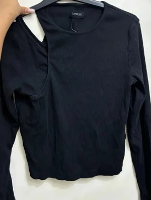 Svart ribbad topp med cut-out från Even&Odd - Säljer en svart långärmad topp från Even&Odd med cool asymmetrisk cut-out vid axeln. Toppen är ribbad och har en tight passform som ger en snygg siluett. Perfekt för dig som gillar trendiga detaljer och vill sticka ut lite extra.