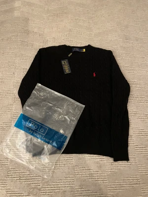 Svart kabelstickad tröja Polo Ralph Lauren - Svart kabelstickad tröja från Polo Ralph Lauren i storlek M. Klassisk rund hals och långärmad modell med röd broderad logga på bröstet. Tröjan är i mjuk bomull och har ribbade muddar vid ärmslut och nederkant. Perfekt för en clean och stilren look.41 cm vid axlarna och 62 cm på längden 