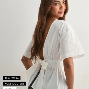 Vit blus med öppen rygg och rosett - Supersöt vit blus med korta ärmar, bred midjedel och stor rosett i ryggen. Blusen har en djup v-ringad öppen rygg och är tillverkad i ett lätt bomullstyg. Perfekt för dig som gillar detaljer och vill ha en feminin look.