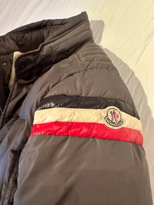 Grå Moncler pufferjacka med detaljer - Säljer en grå pufferjacka från Moncler med hög krage och dragkedja. Jackan har coola detaljer med röd, vit och marinblå rand på ärmen samt Moncler-logga. Fodret är ljusbeige och jackan har två fickor med dragkedja framtill. Perfekt för kalla dagar. ÄKTA SÅ KLART. Passar S/M