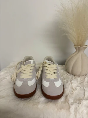 Vita och grå sneakers med brun sula - Snygga sneakers i vitt och ljusgrått med klassisk design. Skorna har en rund tå, platt brun sula och snörning i beige. Materialet är en mix av mocka och syntet, vilket ger en stilren och fräsch look. Perfekta för dig som gillar minimalistisk stil.