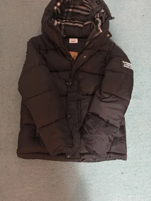 Svart dunjacka från Burberry - Säljer en svart dunjacka från Burberry med huva och klassiskt rutigt foder. Jackan har dragkedja och knappar framtill, samt två fickor med dragkedja. På ärmen sitter en patch med Burberry London England. Perfekt för kalla dagar.