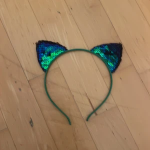 Diadem med kattöron och paljetter - Snyggt diadem med kattöron täckta av glittriga blå och gröna paljetter. Perfekt accessoar för att sticka ut på festen. Diademet är grönt och öronen har en skimrande effekt som fångar ljuset.
