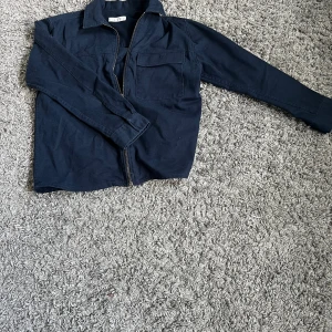 Mörkblå overshirt från Zara - Snygg mörkblå overshirt från Zara med klassisk krage och dragkedja framtill. Jackan har en stor bröstficka och är tillverkad i bomull, vilket gör den både bekväm och lätt att bära. Perfekt för dig som gillar enkel och stilren design.
