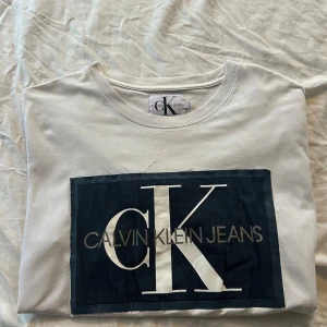 Vit tshirt från Calvin Klein Jeans - Vit T-shirt  från Calvin Klein Jeans med stort svart tryck och CK-logga på bröstet. Rund hals och klassisk passform, tillverkad i mjuk bomull. Perfekt för dig som gillar stilrena och ikoniska märken.