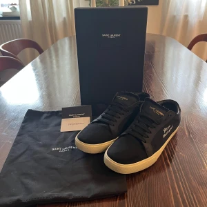 Saint Laurent svarta sneakers - Svarta sneakers från Saint Laurent Paris med vit sula och guldfärgad logga på sidan och plös. Skorna har snörning, rund tå och är tillverkade i canvas med detaljer i skinn. Klassisk och stilren modell med lågt skaft.