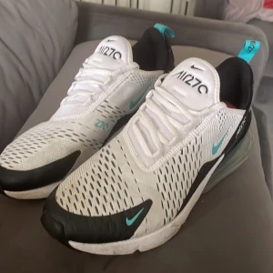 Nike Air Max 270 vita/turkosa sneakers - Nike Air Max 270 sneakers i vitt med svarta och turkosa detaljer. Skorna har en stor synlig Air Max-sula bak, mesh-överdel för bra andning och snörning framtill. Snygg sportig look med turkosa loggor och dragflik bak.