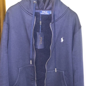Polo Ralph Lauren hoodie - Snygg marinblå hoodie från Polo Ralph Lauren med dragkedja, huva och snörning. Tröjan har klassisk logga broderad på bröstet och stora fickor framtill. Perfekt för en chill och stilren look. Det är storlek M men den sitter som storlek S