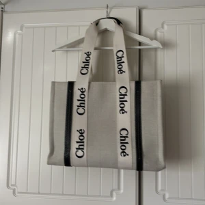 Beige väska - Snygg och rymlig shoppingväska i beige canvas med breda axelband med logotyp. Väskan har svarta detaljer och insida i mörkt tyg. Perfekt för dig som vill ha en trendig och praktisk väska med lyxig känsla. Rymmer en 15 inch Laptop, perfekt till skolan eller jobbet.