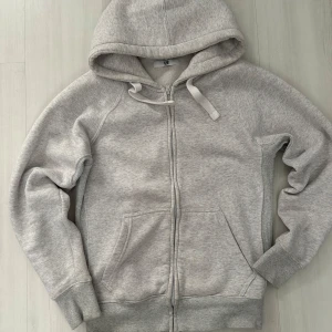 Ljusgrå Russemerch hoodie - Säljer en ljusgrå hoodie från Russemerch i storlek M. Tröjan har huva med snörning, dragkedja framtill och två stora fickor och en liten ficka inne. Mjuk och skön bomullsmix, perfekt för chill dagar. Ribbstickade muddar vid ärmslut och nederkant ger extra komfort.