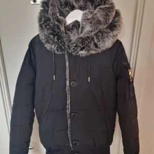 Svart pufferjacka Glorious Gangsta - Svart pufferjacka från Glorious Gangsta med stor fluffig huva i gråmelerad fuskpäls. Jackan har snygga detaljer som guldfärgade snörstoppar, mönstrade fickor, dragkedja på ärmen och knappar framtill. Perfekt för kalla dagar med street vibe.