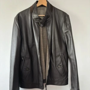 Hackett London jacka - Snygg svart skinnjacka från Hackett med hög krage och dragkedja framtill. Jackan har två fickor med både dragkedja och knappdetalj. Insidan är fodrad med grått rutigt tyg och har en innerficka i skinn med knapp. Perfekt för dig som gillar stilren och klassisk look.