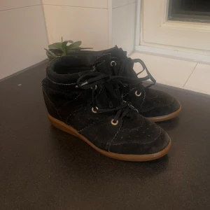 Isabel Marant svarta mockasneakers - Svarta sneakers från Isabel Marant i mjuk mocka med bruna gummisulor. Skorna har snörning med svarta skosnören, dekorativa sömmar och rund tå. Perfekta för dig som gillar en sportig men ändå stilren look. Utbyta skosnören!!!  Ingen låda till skorna!