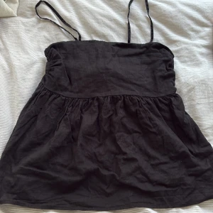 Svart topp från Lindex - Svart topp från Lindex med smala axelband och  kjol. Överdelen har smock baktill för en bekväm passform. Tillverkad i ett lätt bomullstyg, perfekt för varma dagar. Enkel och stilren design som är lätt att matcha. Toppen är aldrig använd och i storlek S.