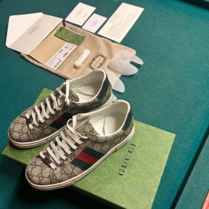 Gucci Ace sneakers - Gucci Ace sneakers i beige canvas med klassiskt GG-monogram, gröna och röda sidoremsor och vita skosnören. Skorna har rund tå, platt sula och vita detaljer vid häl och sula. Kommer med originalkartong, extra skosnören och tillbehör. Går att pruta på priset. Det är bara att skriva