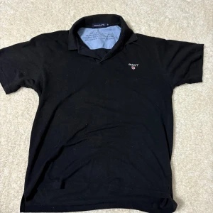 Svart pikétröja från GANT - Klassisk svart pikétröja från GANT med liten broderad logga på bröstet. Tröjan har krage, korta ärmar och är tillverkad i mjuk bomull. Perfekt för en clean och stilren look.