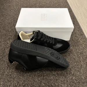 Svarta sneakers från Maison Margiela - Snygga svarta sneakers från Maison Margiela med ovandel i skinn och mocka. Skorna har svarta snören, diskret logga på plösen och en klassisk rund tå. Insidan är fodrad i ljus textil för extra komfort. Perfekta för dig som gillar stilrena och exklusiva sneakers.