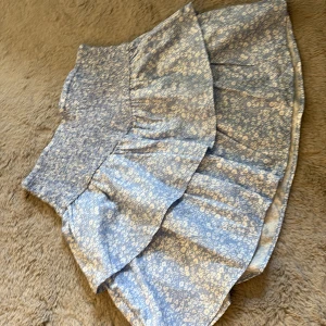 Blommig blå volangkjol från Gina Tricot - Supersöt blå kortkjol från Gina Tricot med vita små blommor och volangdetaljer. Kjolen har bred smock i midjan för en bekväm passform och är perfekt för en fräsch och somrig look.