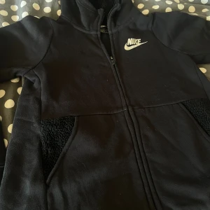 Svart fleecejacka från Nike - Svart fleecejacka från Nike med dragkedja och hög krage. Jackan har mjukt teddyfoder på insidan och i fickorna, samt klassisk Nike-logga på bröstet. 