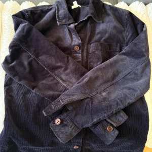 Svart manchester overshirt från H&M - Säljer en mörkblå overshirt i manchester från H&M. Skjortan har klassisk krage, bröstficka och knäppning framtill med bruna knappar. Perfekt att slänga över en t-shirt för en chill look. Långärmad och i mjukt, strukturerat tyg.