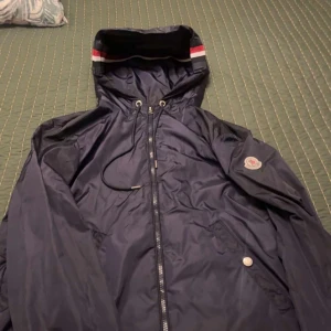 Mörkblå vindjacka från Moncler - Snygg mörkblå vindjacka från Moncler med huva och dragkedja framtill. Jackan har vita och röda detaljer på huvan, snörning vid halsen och Moncler-logga på ärmen. Perfekt för dig som vill ha en stilren och sportig look.