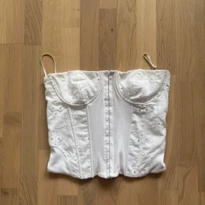 Vit broderad korsett från Zara - Supersnygg vit korsett från Zara med tunna axelband och broderade detaljer. Toppen har markerade kupor och en figurnära passform som ger en riktigt trendig siluett. 