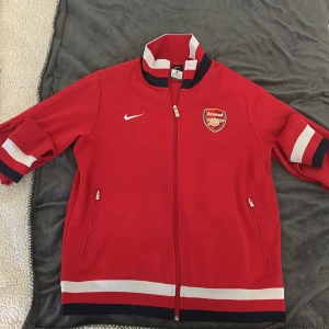 Röd Arsenal Nike fotbollströja - Snygg röd Arsenal fotbollströja från Nike med dragkedja, broderat klubbmärke och vita samt mörkblå detaljer på krage, ärmslut och nederkant. Tröjan har två fickor med dragkedja och tydlig Arsenal-text på ryggen. Perfekt för dig som älskar fotboll och vill sticka ut.
